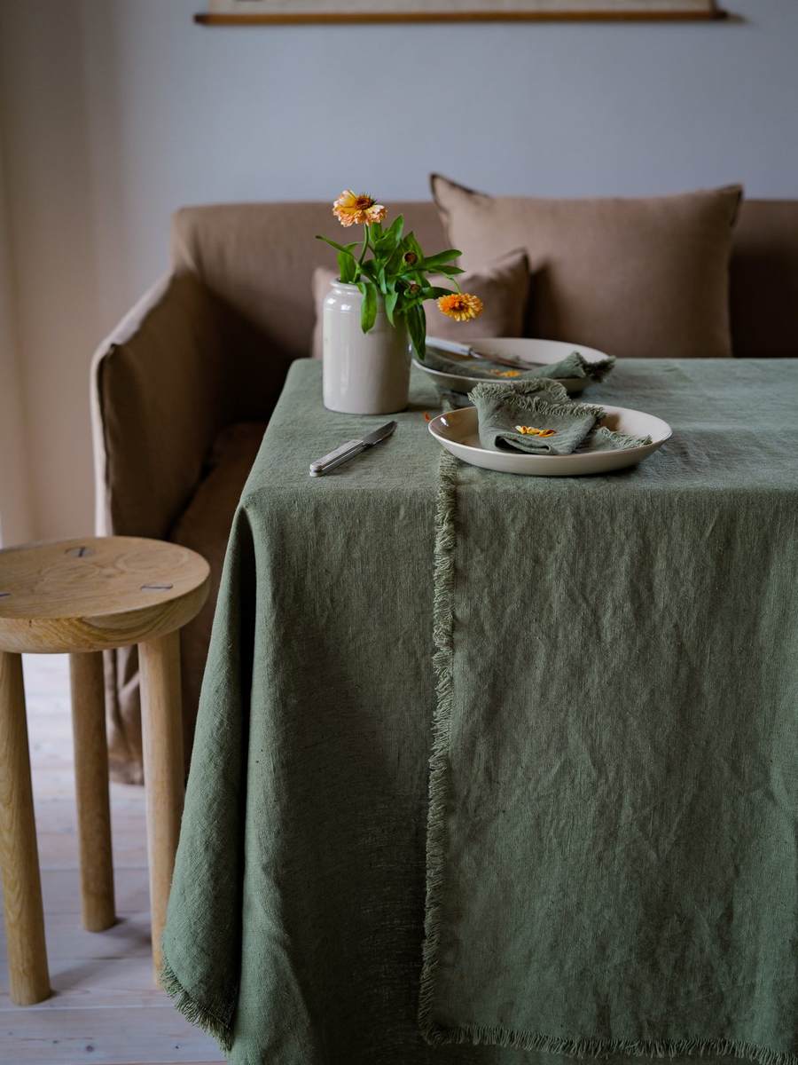 Mantel de lino verde oliva con flecos, platos crema y jarrón de flores